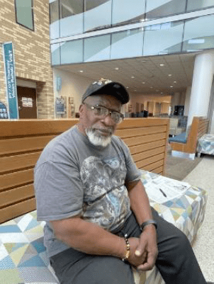 An older man with a beard sits on a patterned bench in a modern atrium. He wears a graphic T-shirt and cap.