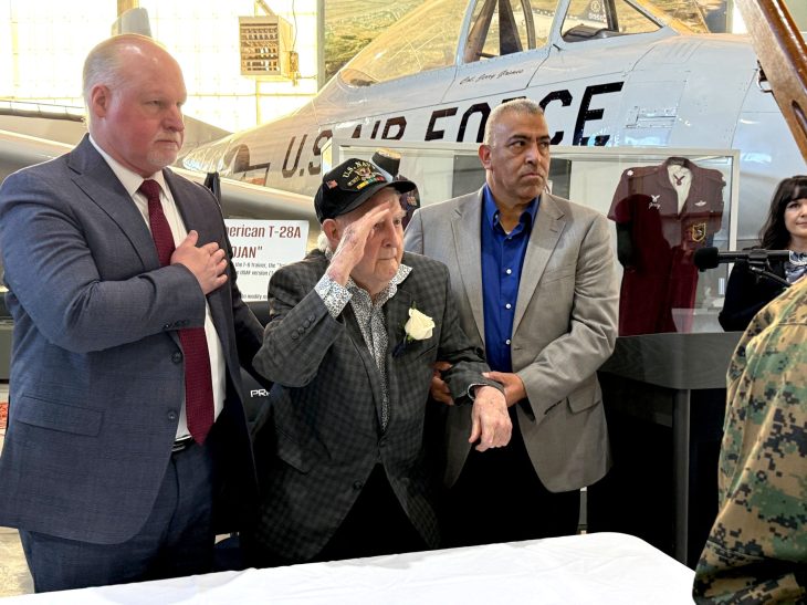 An elderly veteran in a military cap salutes, supported by two men, in front of a vintage U.S. Air Force plane at a museum. The atmosphere is respectful.