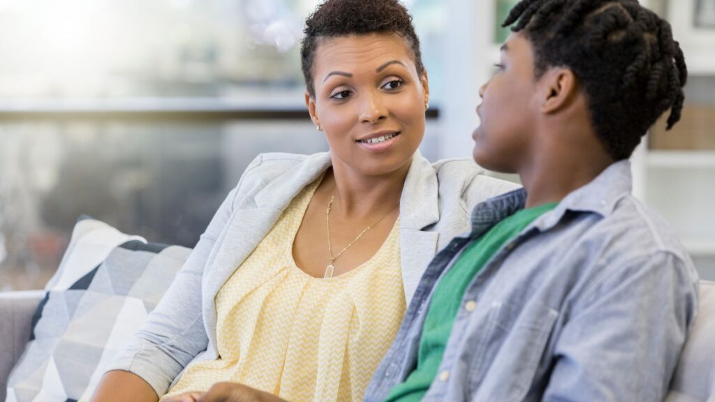 A mother and teenage child sitting on a couch having a thoughtful conversation, making eye contact in a bright living room.